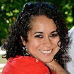 A person with curly hair, wearing earrings and a red top, is smiling against a blurred leafy background.