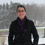 A person in a black coat and glasses smiles while standing outdoors in a snowy landscape.