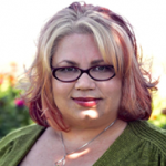 Person with glasses and multicolored hair, wearing a green top, standing outdoors with a blurred background of flowers.