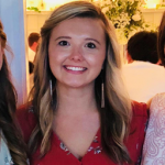 Smiling woman with long blonde hair wearing a red patterned dress is standing indoors.