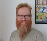 A man with a long red beard and glasses is smiling, standing in front of a framed artwork depicting cars.