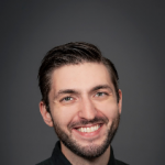 Smiling man with short dark hair and a beard, wearing a black button-up shirt, against a dark gray background.