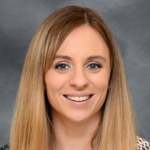 A woman with long, straight blonde hair smiles at the camera. She is wearing a patterned top and a necklace. The background is a plain, gradient gray.