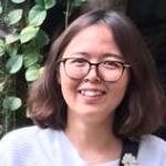 A person with shoulder-length brown hair and glasses smiles, standing in front of leafy greenery, wearing a light-colored top.