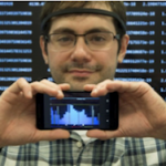 Man with glasses holds a smartphone displaying a graph, wearing a headband. A screen with data in the background.