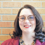 A person with glasses is smiling in front of a brick wall, wearing a maroon jacket and a beaded necklace.