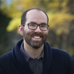 Smiling man with a beard, glasses, and a blue sweater stands outdoors with blurred autumn leaves in the background.