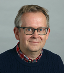 A person with glasses and a blue sweater over a red and white checked shirt smiles at the camera.