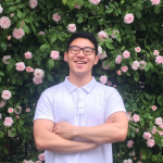 Person wearing glasses and a white shirt stands with arms crossed, smiling in front of a bush with pink flowers.