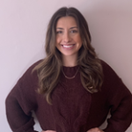 A person with long brown hair wearing a dark sweater stands smiling against a plain light-colored wall.