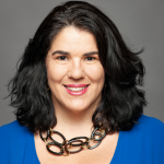 A person with shoulder-length dark hair, wearing a blue top and a chunky necklace, smiles against a gray background.