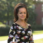 A woman with dark hair wearing a floral dress and gold necklace stands outdoors in a park-like setting.