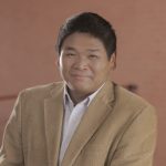 A person in a corduroy blazer and striped shirt poses with a pleasant expression, standing by a railing indoors with a blurred background.