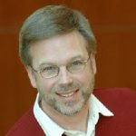 A person with glasses, a beard, and short hair smiles, wearing a red sweater over a white shirt.