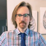 Man with glasses and a beard wearing a checkered shirt and blue tie stands indoors with artwork on the wall behind him.