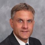 A person wearing a striped suit and tie against a gray background.