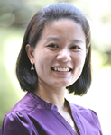 A woman smiling, wearing a purple blouse and earrings, standing outdoors with a blurred background.