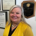 A person with long gray hair smiles in front of framed certificates and an award plaque on a wall. They are wearing a yellow jacket and necklace.