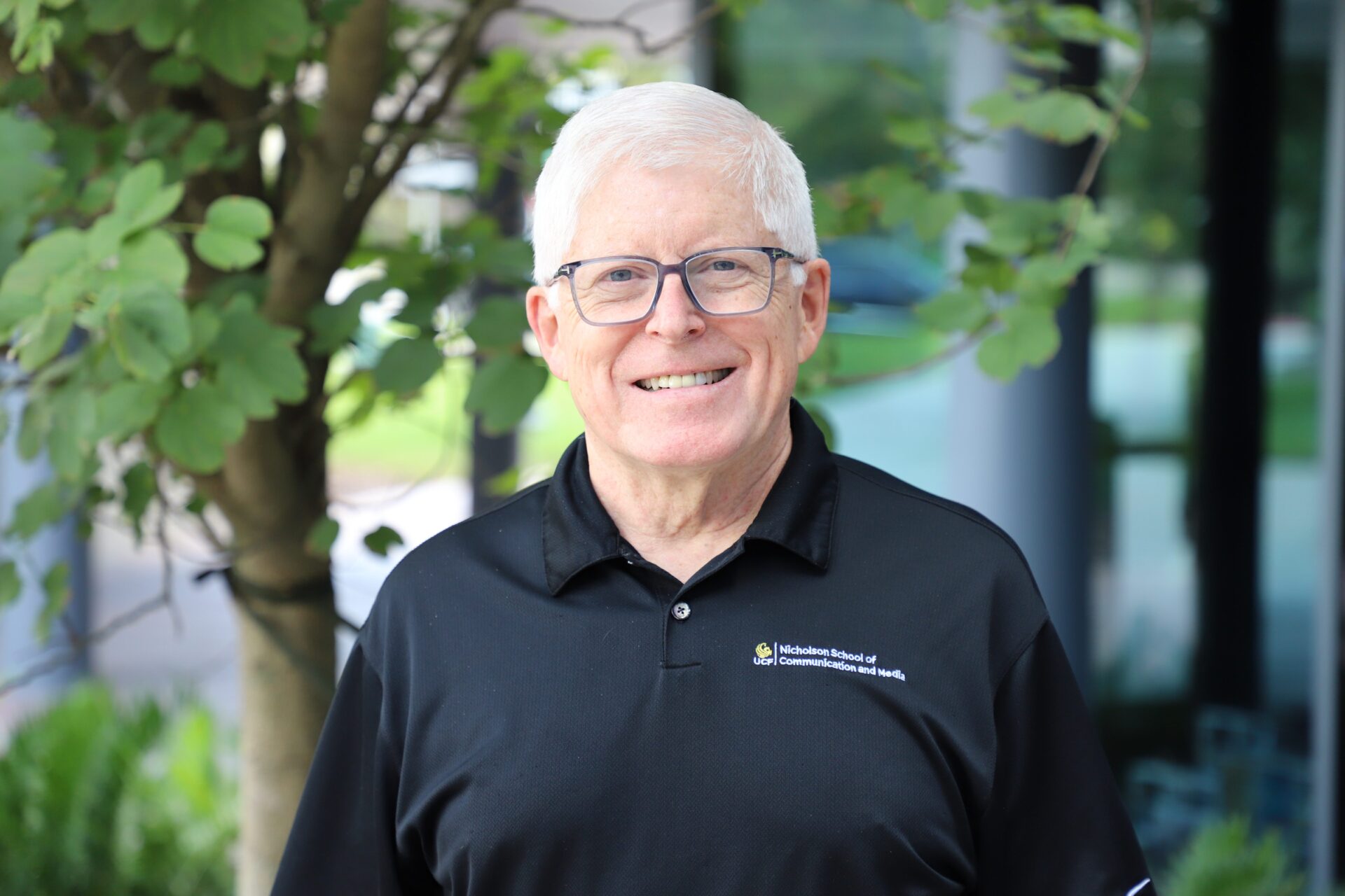 Older man with short white hair and glasses, wearing a black collared shirt, smiling outdoors near a tree with green leaves.