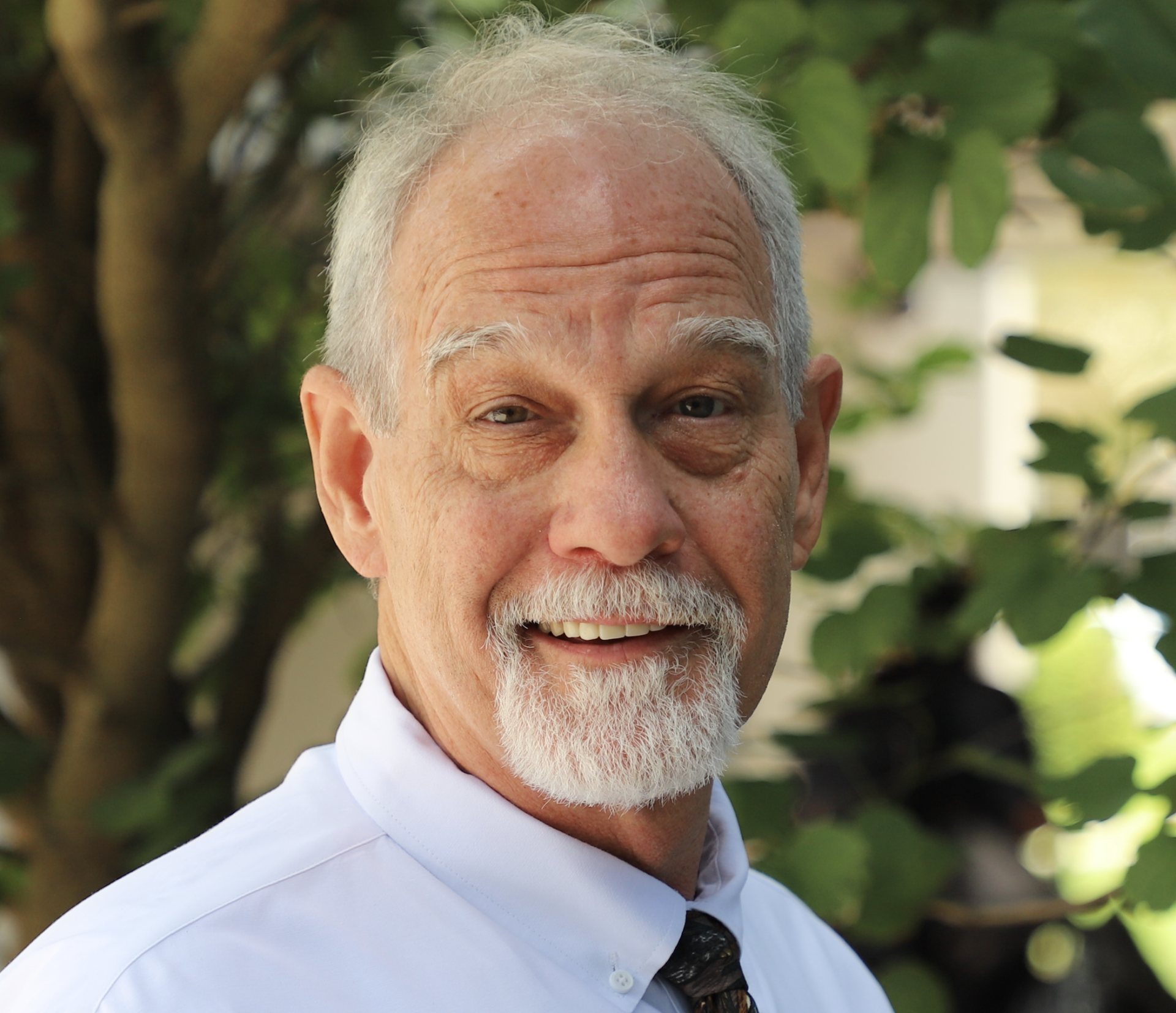 An older man with white hair and a beard, wearing a white shirt and tie, stands outdoors in front of green foliage, smiling at the camera.