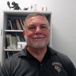 A man with a beard wearing a "Knights UCF" shirt sits in front of a bookshelf, smiling at the camera.