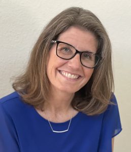 A woman with shoulder-length brown hair and glasses smiles while wearing a blue top.