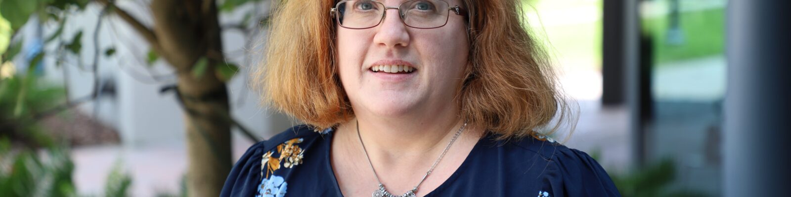 A woman with glasses and shoulder-length red hair stands outdoors near a tree, wearing a navy blue floral blouse.