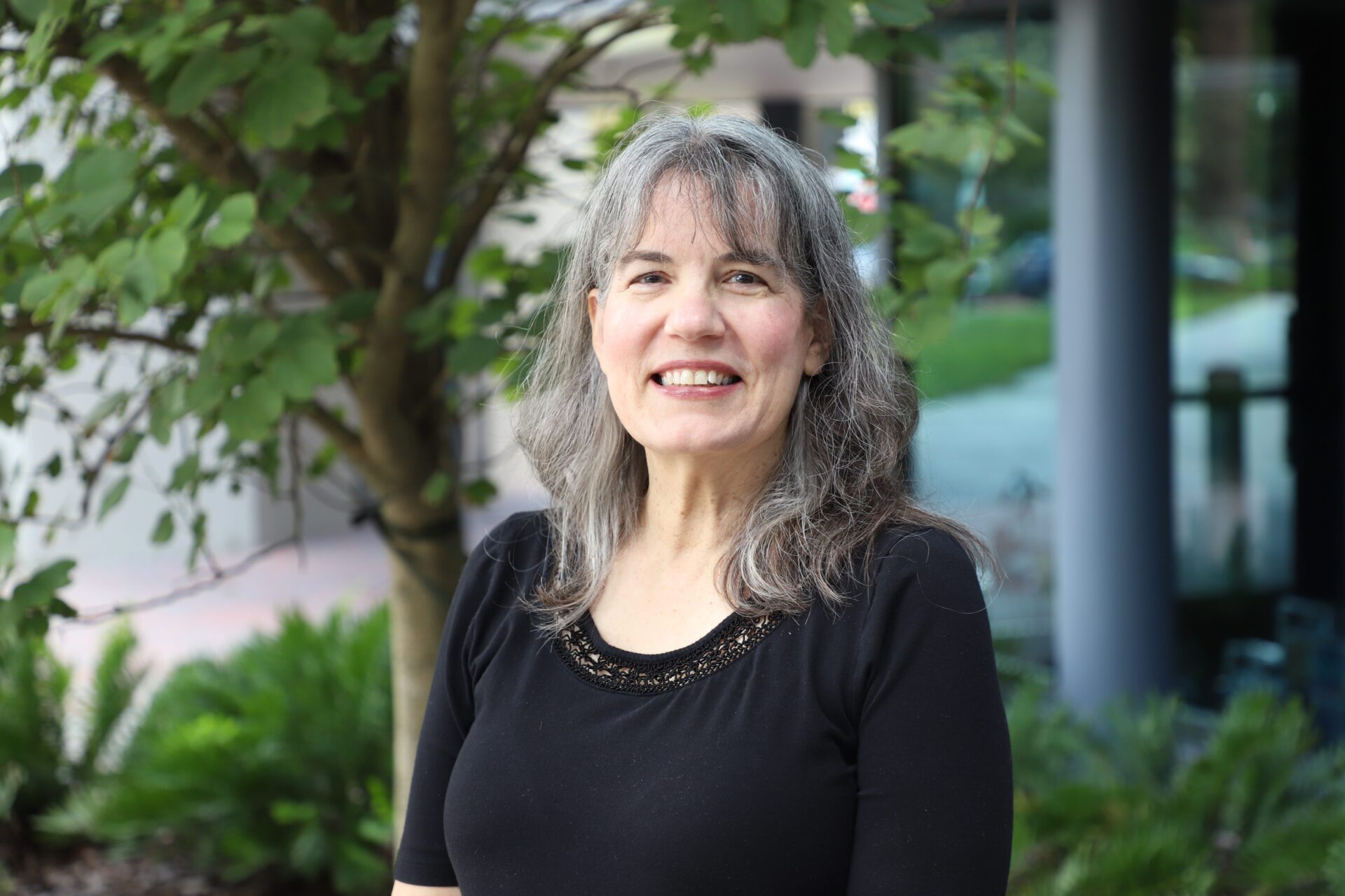 A woman with gray hair wearing a black top stands outside near a tree and greenery, smiling at the camera.