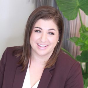 Woman with brown hair wearing a maroon blazer, smiling in front of a green plant.