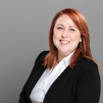 A person with red hair and a black suit jacket smiling against a gray background.