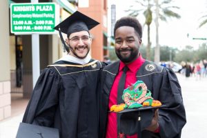 Two graduates in black caps and gowns stand together. One holds a decorated cap with a dragon design. A parking sign and palm trees are in the background.