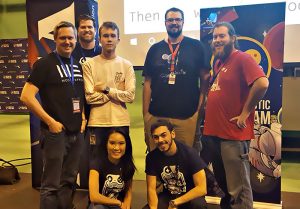 A group of seven people posing together in front of promotional banners at an event. Some are standing, and some kneel in front.