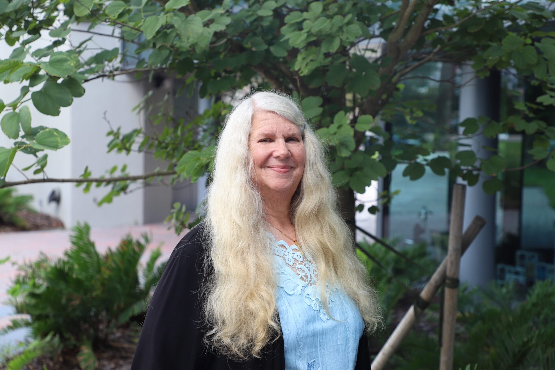 Woman with long blonde hair stands outdoors in front of a leafy tree, wearing a light blue blouse and a black jacket, with greenery and a building in the background.