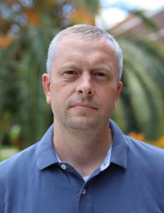 A man with short gray hair and a neutral expression, wearing a blue polo shirt, stands outside with a palm tree in the background.