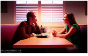 A man and woman sit across from each other at a table in a dimly lit restaurant, engaged in conversation.