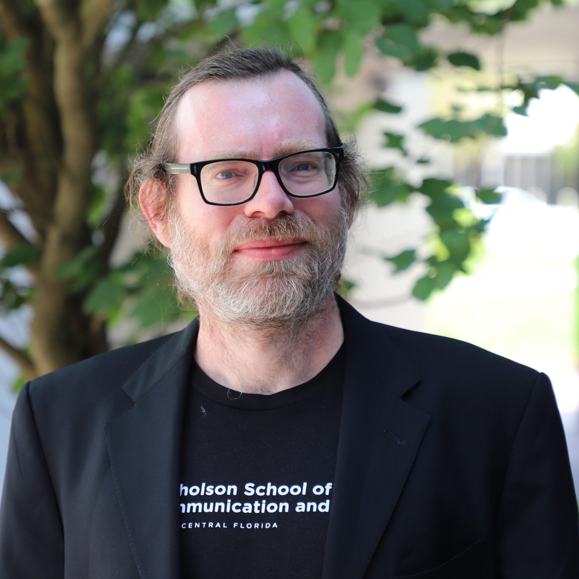A man with glasses, a beard, and a black jacket stands outdoors in front of a leafy background, wearing a “Nicholson School of Communication and Media, Central Florida” T-shirt.
