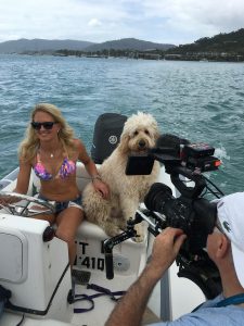 Person in a colorful bikini sits on a boat with a large fluffy dog, while a cameraman films them. Water and distant hills are in the background.