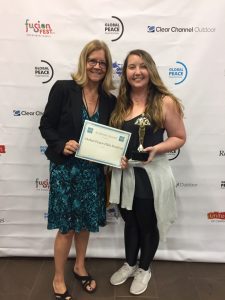 Two women standing in front of a branded backdrop. One holds a certificate labeled "Global Peace Film Festival," and the other holds a trophy.