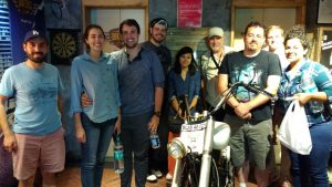 A group of people posing indoors, standing behind a motorcycle. Some are holding water bottles. There is a dartboard and posters in the background.