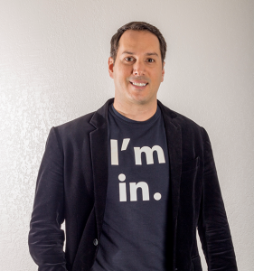 A person stands against a white wall, wearing a dark blazer and a T-shirt with the phrase "I'm in.