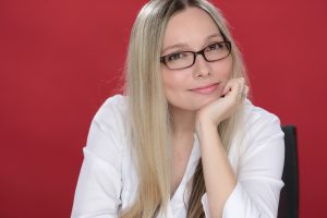 A person with long blonde hair and glasses, wearing a white shirt, sits with their chin resting on one hand against a red background.