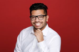 Man wearing glasses and a white shirt smiles against a red background, resting his chin on his hand.