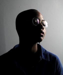 A person with short hair and glasses is partially lit against a dark background, eyes closed and wearing a dark shirt.