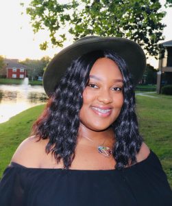 A person wearing a hat and off-shoulder top smiles in an outdoor setting near a pond and trees.