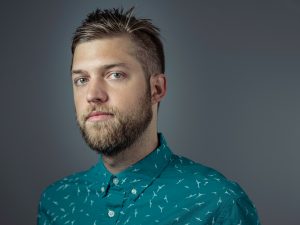 A man with a beard and short hair wearing a teal shirt with white patterns, looking directly at the camera.