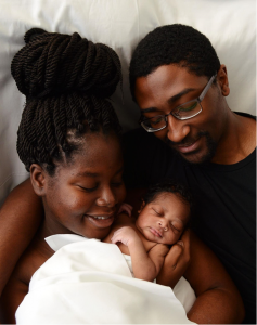 A couple lovingly holds their newborn, wrapped in a white blanket, while lying on a bed.