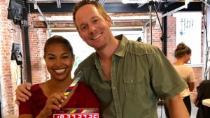 Two people smiling and holding a clapperboard in a film set with brick walls in the background.