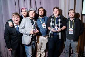 Six men standing together, three holding awards, at an event with "GDC" lanyards.