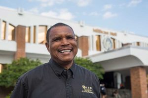 Smiling man in a black UCF College of Arts and Humanities shirt stands outdoors in front of a building.