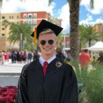 A graduate wearing a cap and gown smiles for a photo outdoors, with palm trees and a crowd in the background.